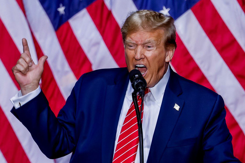 Donald Trump speaking into a microphone and raises his hand in front of an American flag