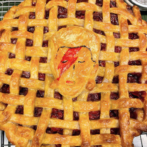 an overhead photo of a baked cherry pie with lattice crust and a dough cutout of a man with a red lightning bolt across his face