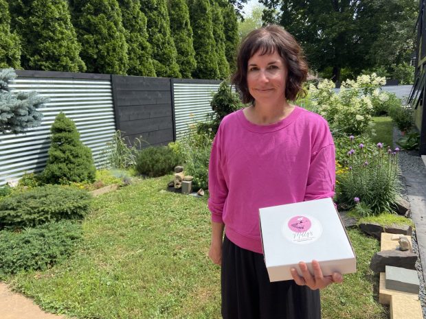 Sarah Millfelt stands in a manicured yard holding a white box with a pink sticker on it.