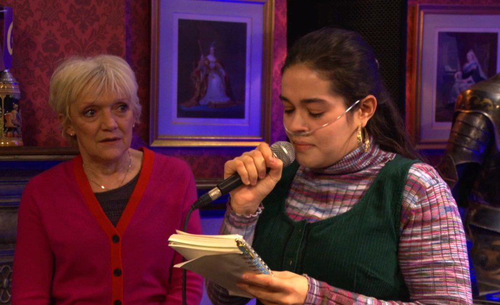 Jean watching Jade speak into a microphone while reading from her notebook in EastEnders