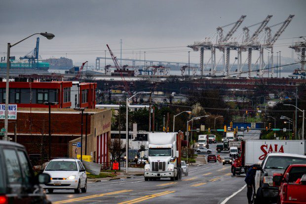 Mar 27, 2024: Idled cranes at the Port of Baltimore and collapsed bridge are seen in the distance as trucks move through the intersection of Broeining Highway and Holabird Avenue. (Jerry Jackson/Staff)